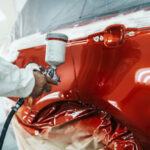 Man With Protective Clothes And Mask Painting Car Using Spray Compressor.